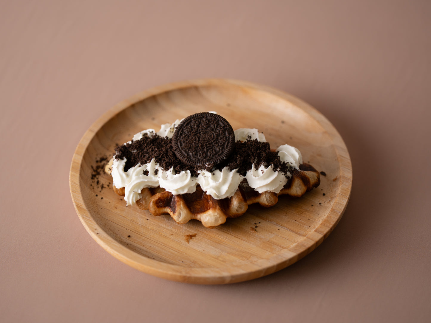 Waffle with whipped cream and Oreo cookies on a wooden plate against a brown background