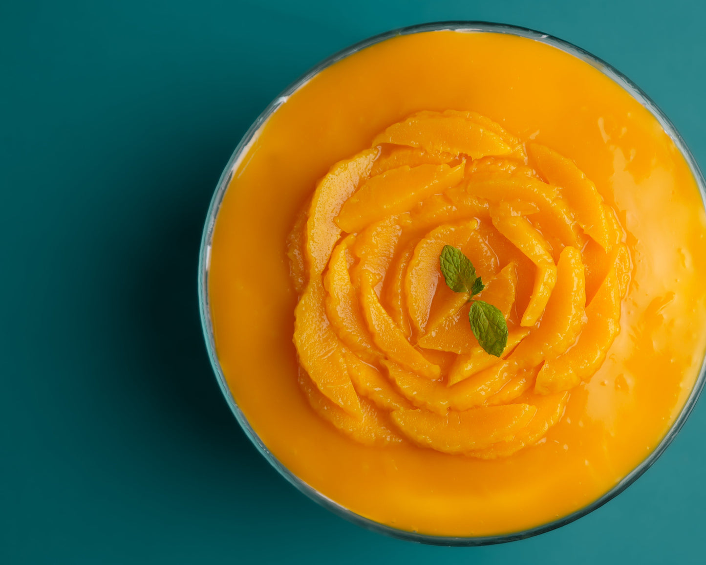 A top view of a Mango Trifle dessert with layers of cake and cream, garnished with a mango rose on top, on a teal background.